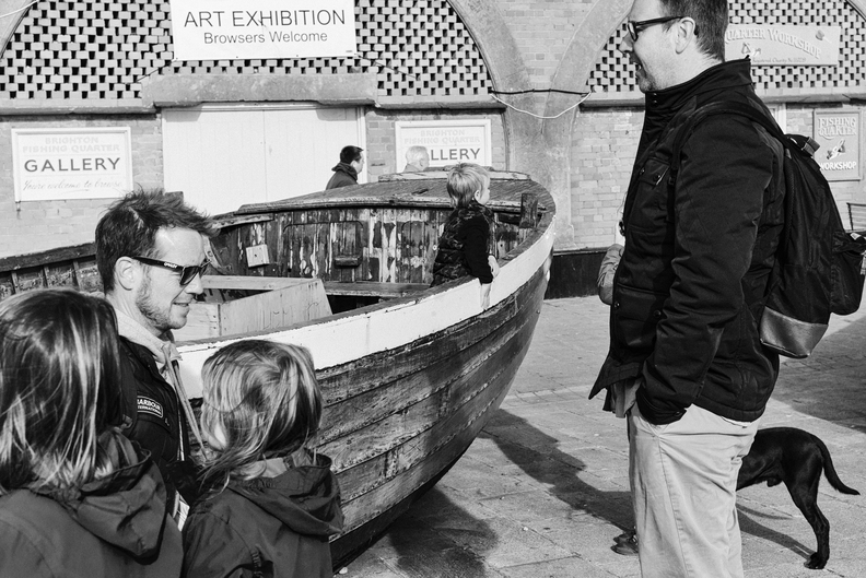 Photo by Heathcote called Browsers Welcome shows a wooden boat, a cluster of people, signs at the back saying Art Exhibition: browsers welcome