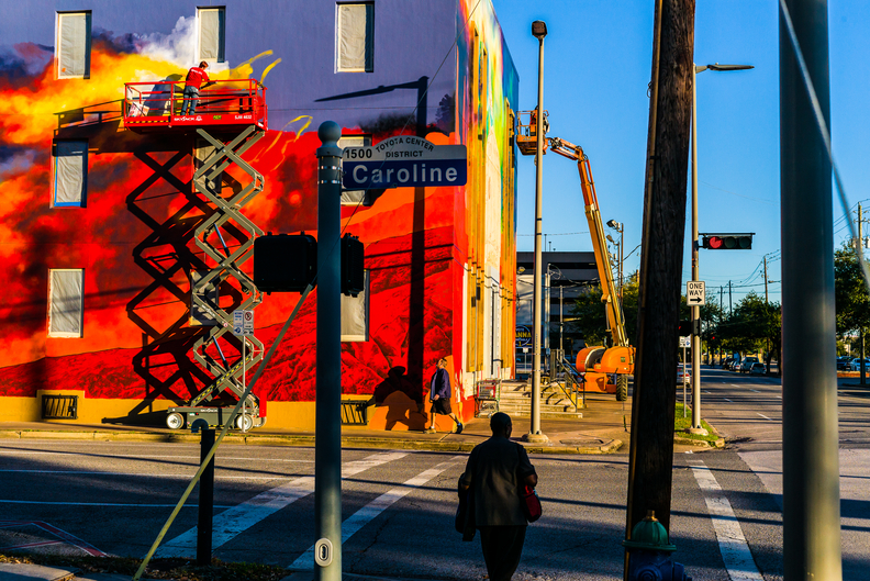Photo by Heathcote called Caroline shows bright colours and harsh light on a street corner in Houston, maybe a building site, a bright red wall and a road sign saying Caroline