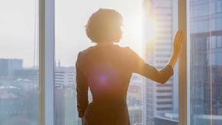 Shot of the Successful Businesswoman in a Striking Black Dress in Her Office Looking out of the Window at Sunset. Modern Business Office with Personal Computer and Big City View