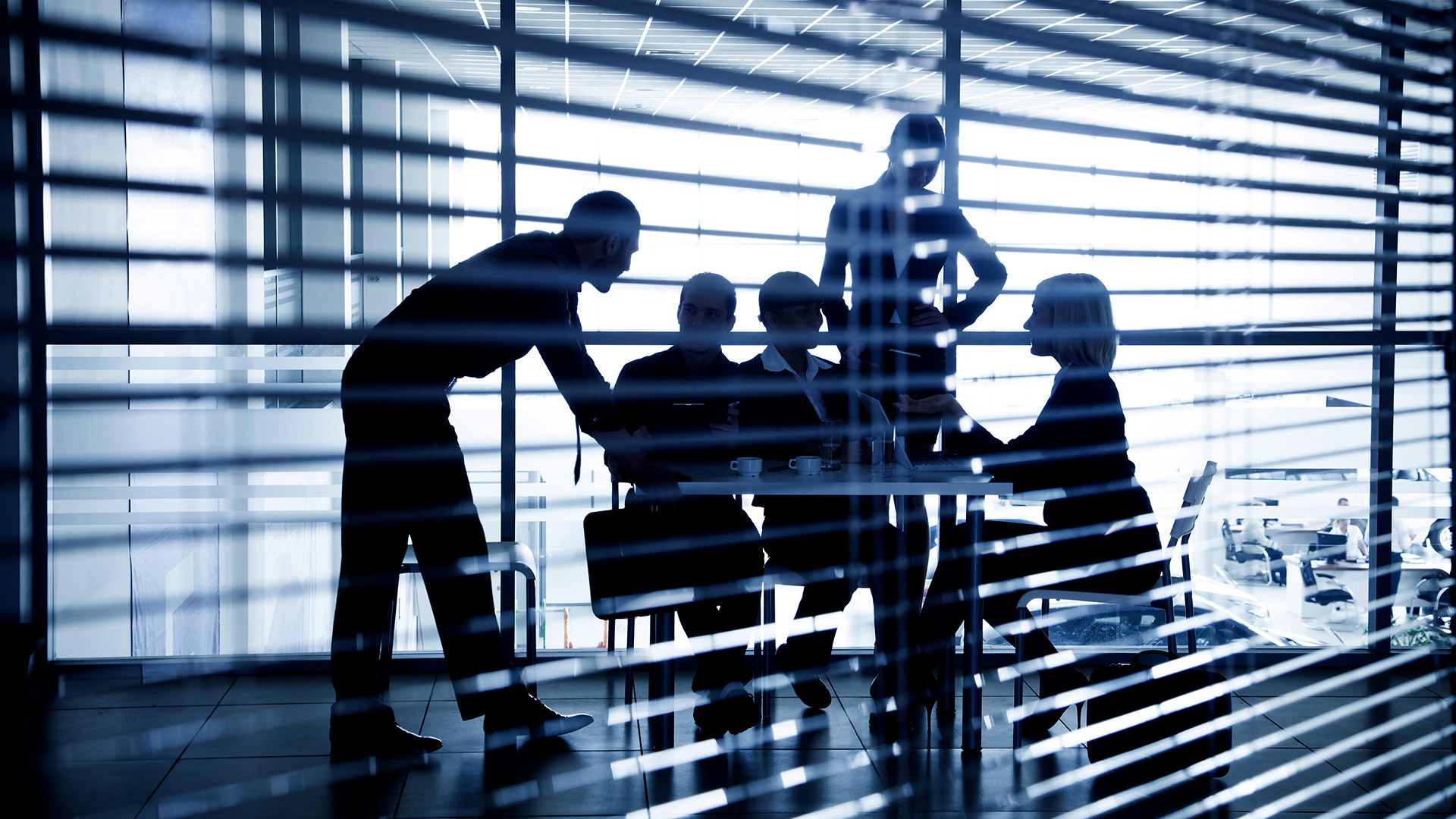 Looking through venetian blinds into boardroom, five people are having a meeting