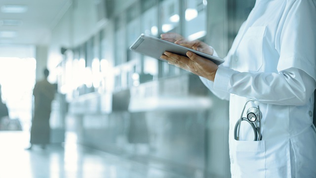 A doctor in a whitecoat holding a tablet in a hospital hallway