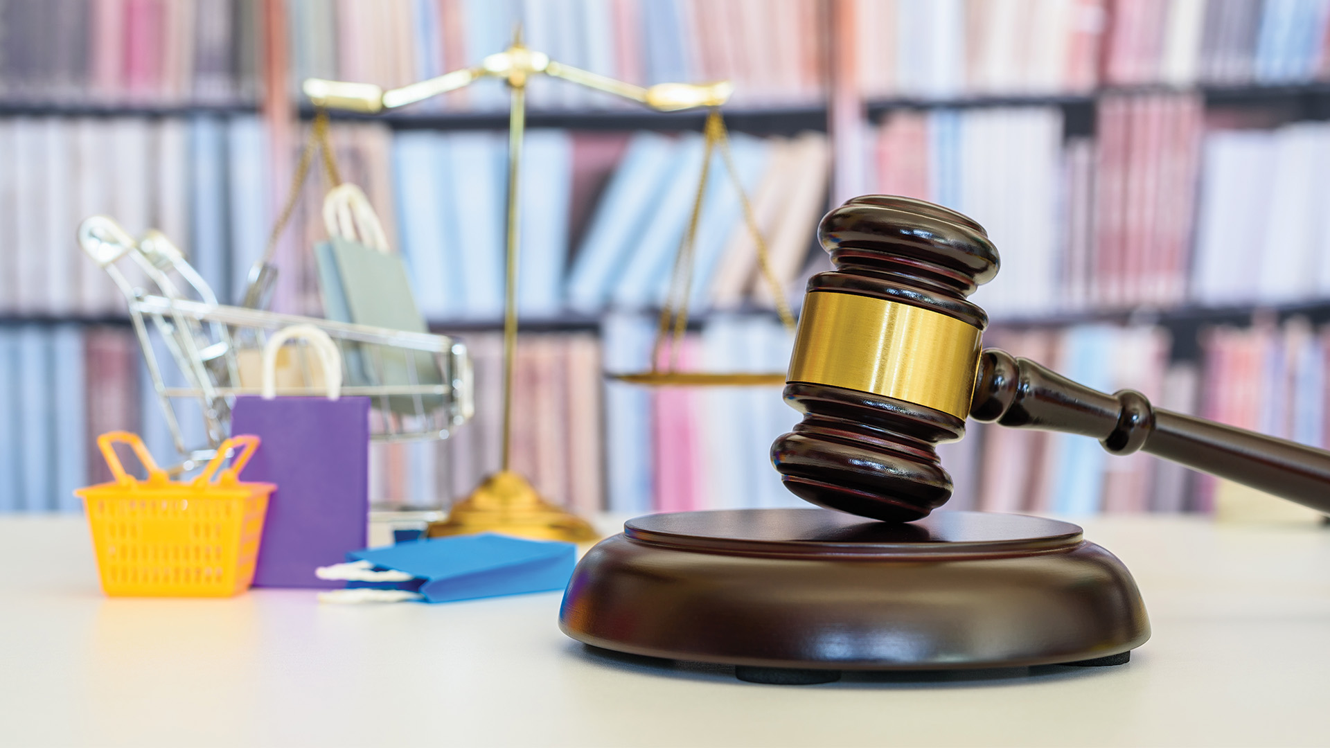 Gavel, scales of justice and visual representation of the consumer products industry of colored shopping bags on table in front of wall of books