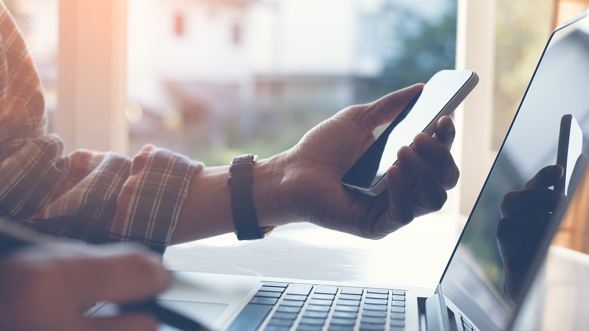 Close up of casual man or freelancer working on laptop computer and holding mobile smart phone with reflection on blank screen